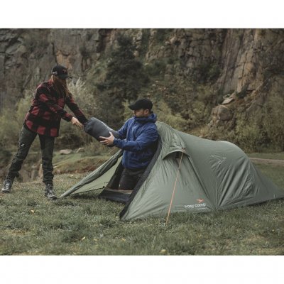 Reindal 2 tunnel tent in a natural environment, two people carry a sleeping bag through the tent's open entrance.